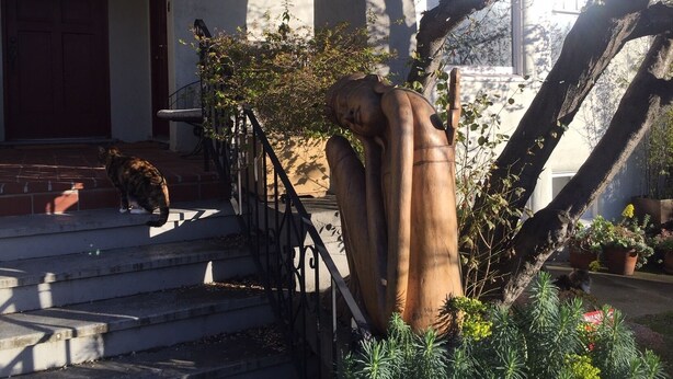 Eine Treppe führt zu einem Haus, im Vordergrund steht eine große Holzskulptur.