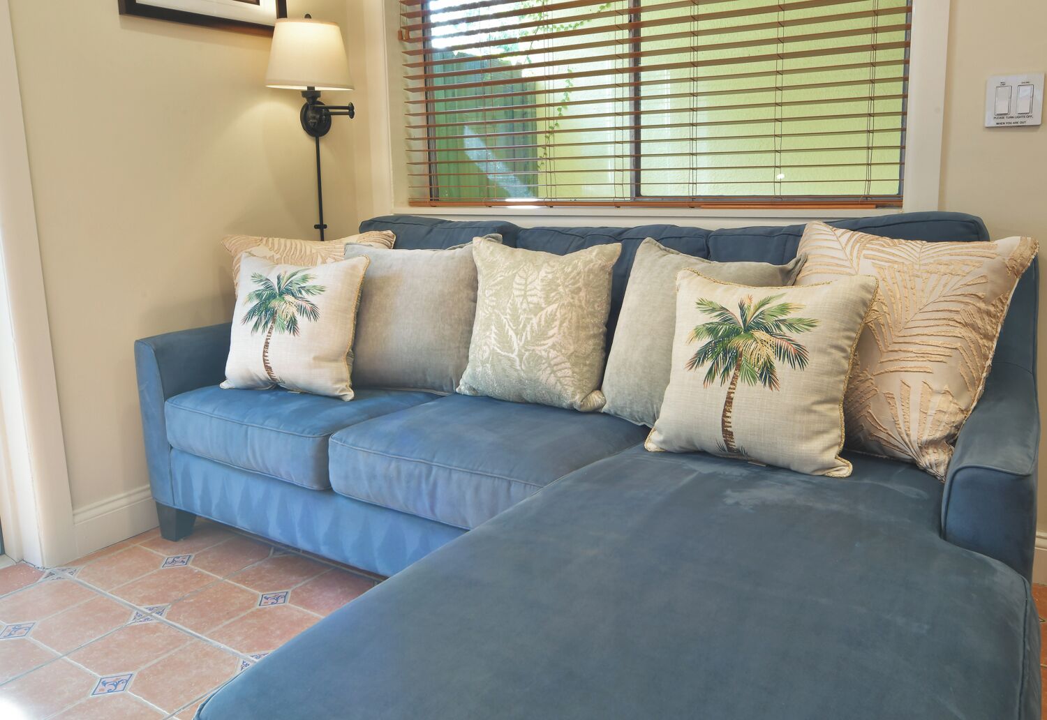 A blue sofa with palm tree and leaf patterned cushions, a floor lamp, and a window with blinds.