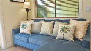 A blue sofa with palm tree and leaf patterned cushions, a floor lamp, and a window with blinds. - Walk to GAY Wilton Drive, Pool, Queen Bed, Kitchen, Living Room, Patio, WiFi, #3 (Wilton Manors)