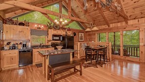 Ein rustikales Blockhaus-Interieur mit Holzbalken, einer Küche mit modernen Geräten, einem Essbereich mit einer Bank und großen Fenstern mit Blick ins Freie.