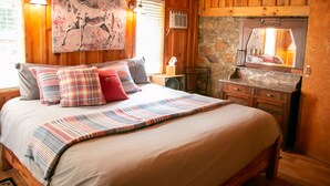 A wooden cabin-style bedroom with a large bed, plaid bedding, a mounted painting, and a stone fireplace. - Victory Suite - Rustic Cabin (Hot Springs)