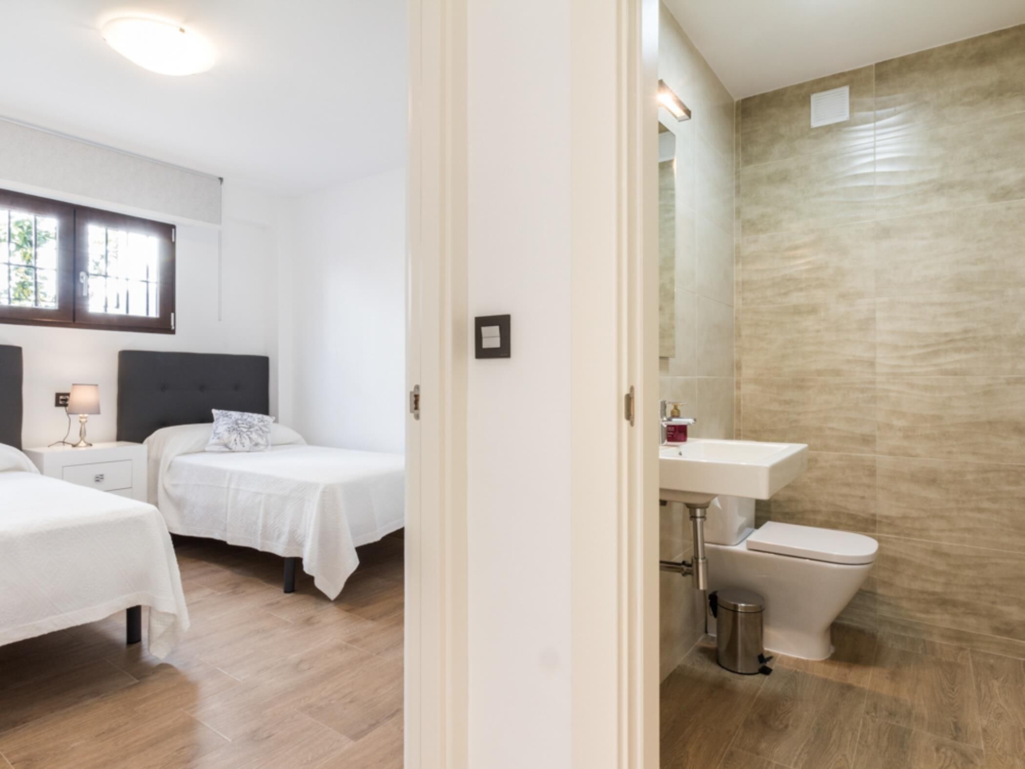 A hotel room with two beds, a bathroom with a sink and toilet, and a window with blinds.