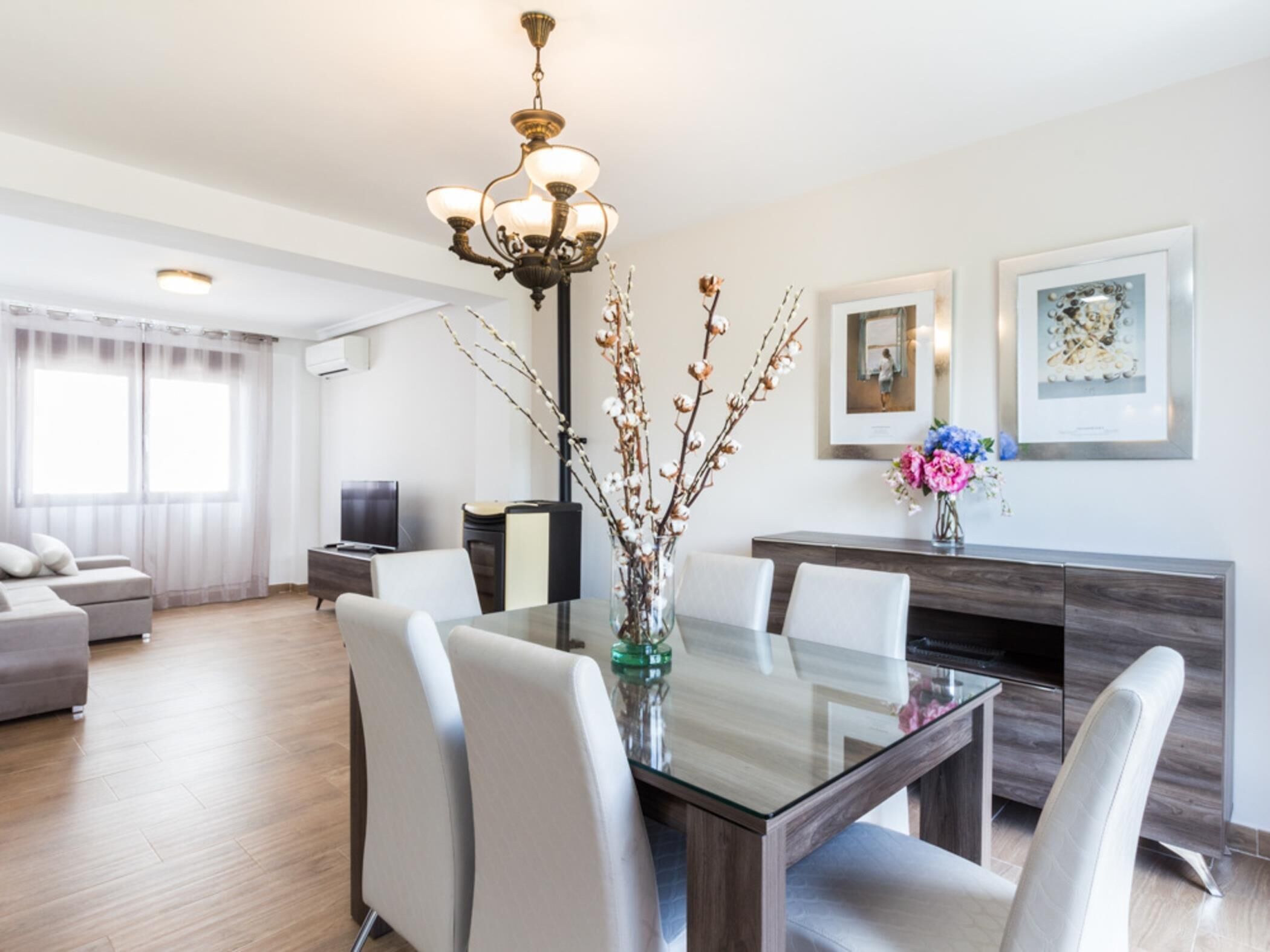A modern living room with a glass dining table, white chairs, a chandelier, and a sofa.