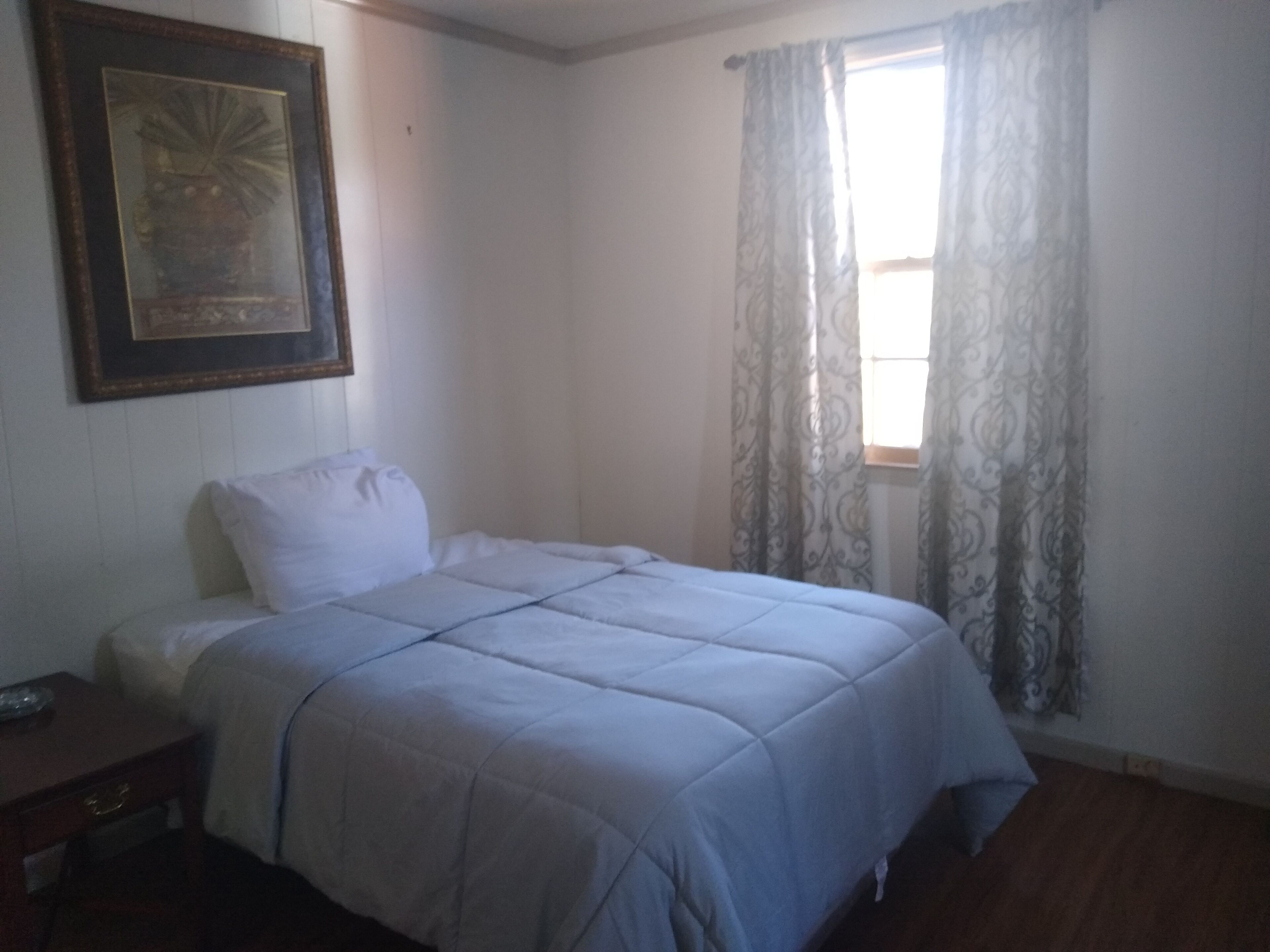Une chambre à coucher avec un lit, une table de chevet, un tableau encadré au mur et une fenêtre avec des rideaux.