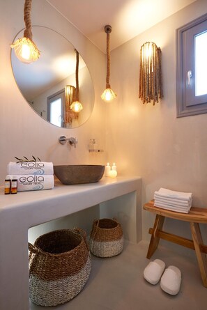 A bathroom with a round mirror, a large vessel sink, and a wooden shelf with towels. There are woven baskets on the floor and a wooden stool with slippers. - Traditional Villa with Private Heated Pool (Santorini)