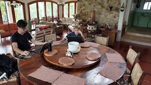 Une table de salle à manger ronde en bois, avec une tasse blanche et un thermos noir. Des sets de table en fibres tressées et des chaises sont disposés autour de la table.