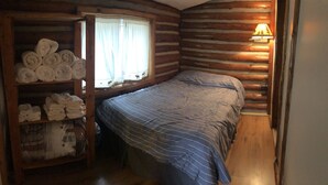 A cabin-style room with a bed, a shelf with towels, a window with curtains, and a wall-mounted lamp. - Cabin 6 Lake Cabins Resort on Fife Lake (Fife Lake)