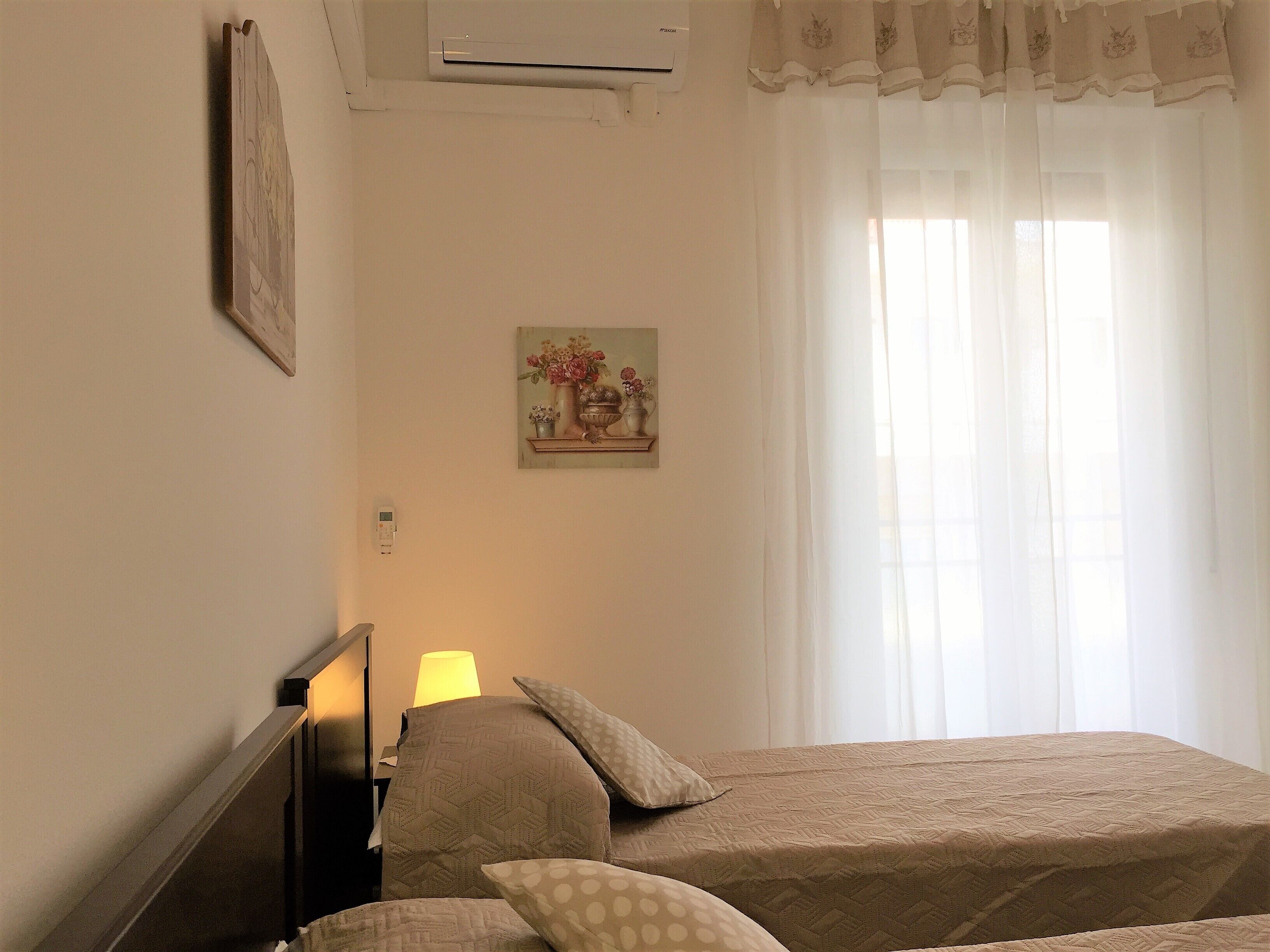A bedroom with two beds, a nightstand, a lamp, a painting on the wall, and a window with curtains.