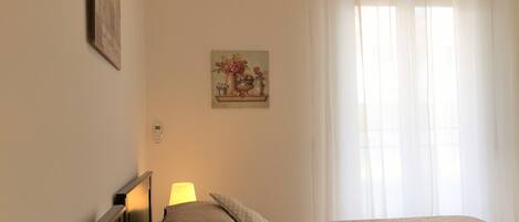 A bedroom with two beds, a nightstand, a lamp, a painting on the wall, and a window with curtains.