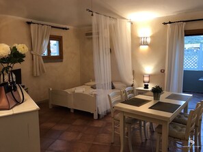 A bedroom with a bed, bedside table, and a small table with chairs. There is a window with curtains, a wall-mounted lamp, and a view of the outside.