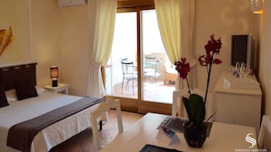 A hotel room with a bed, a desk with a computer, a chair, a small table with a vase of flowers, and a view of the outside balcony.