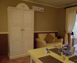 A room with a white wardrobe, a beige sofa, a table with a placemat and a vase of flowers, and an air conditioning unit on the wall.