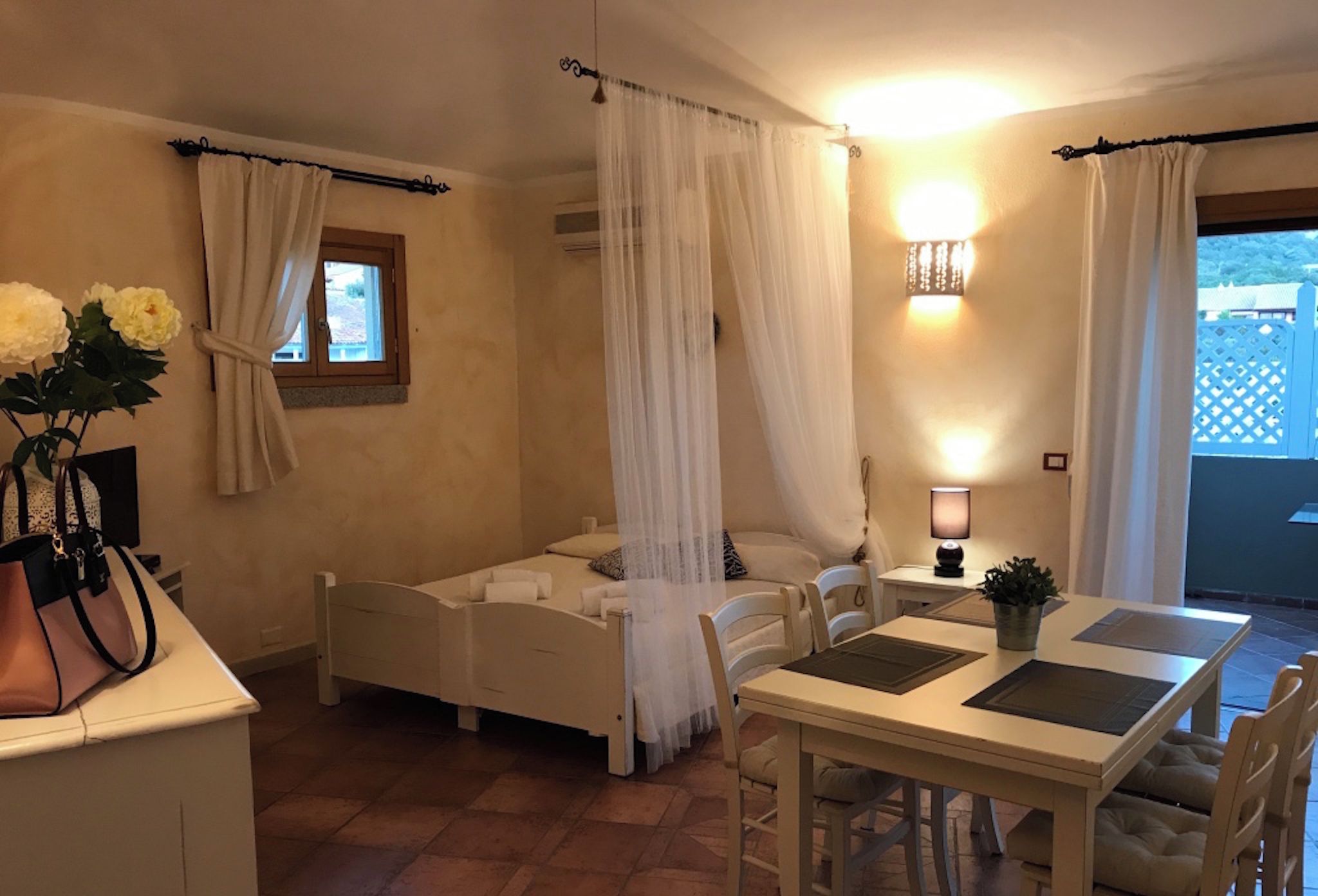 Un dormitorio con cama, mesita de noche y una pequeña mesa con sillas. Se ve una ventana con cortinas, una lámpara de pared y vistas al exterior.