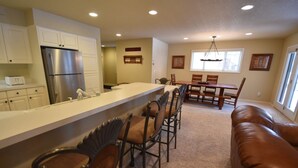 A modern kitchen with a stainless steel refrigerator, a white countertop, and a dining area with wooden chairs and a brown leather couch. - Mountain Harbor #163 3 Bedroom Condo (Whitefish)
