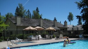 A swimming pool with lounge chairs and umbrellas, surrounded by a fence and trees. - Mountain Harbor #163 3 Bedroom Condo (Whitefish)
