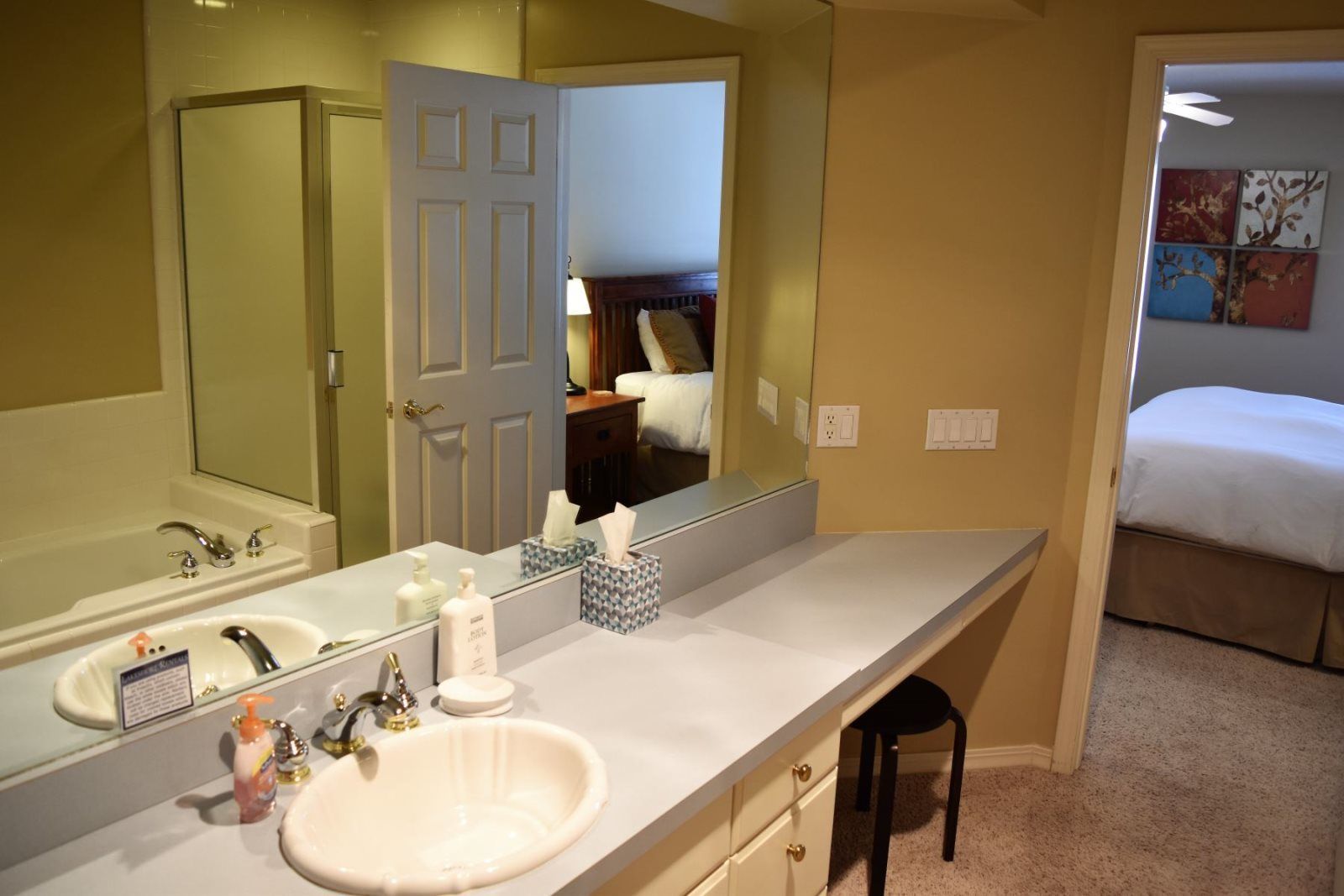A hotel bathroom with a large mirror, a sink, and a toilet. There is a bed visible through the door in the adjacent room.