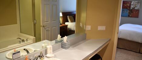 A hotel bathroom with a large mirror, a sink, and a toilet. There is a bed visible through the door in the adjacent room.