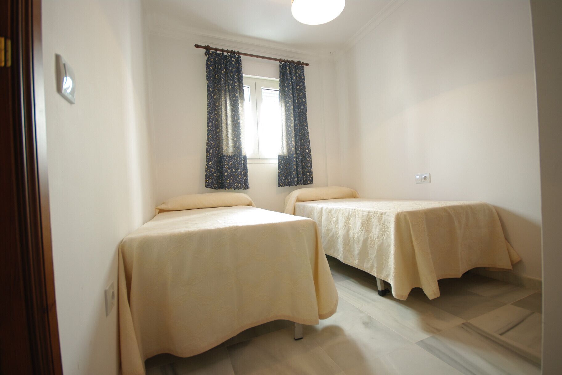 Une chambre avec deux lits simples, une fenêtre ornée de rideaux à motifs et un interrupteur sur le mur.
