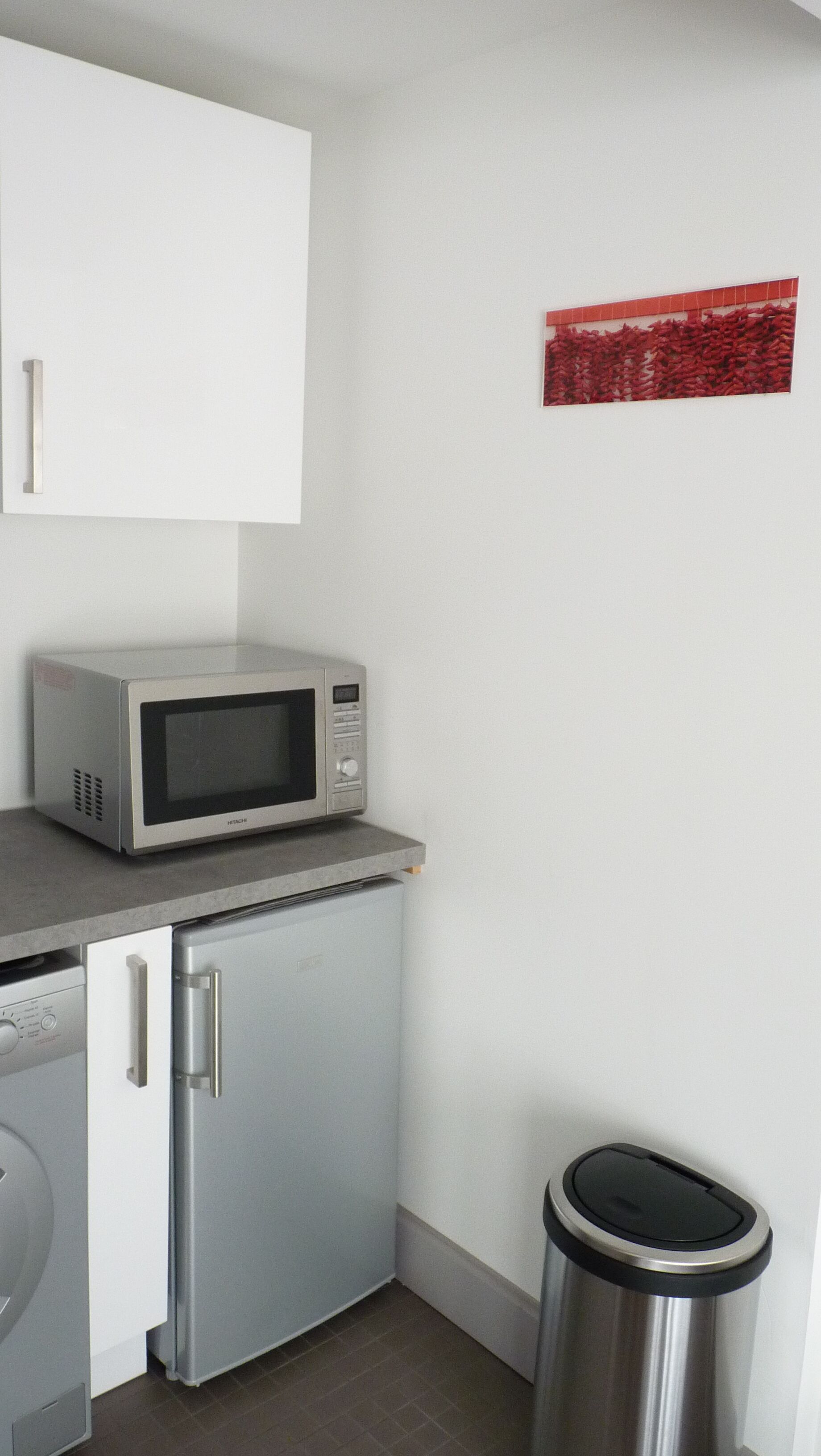 Eine kompakte Küchenzeile mit Mikrowelle, kleinem Kühlschrank, Waschmaschine und Mülleimer.