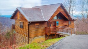 A log cabin with a stone foundation, a wooden deck, and a scenic overlook. - 🌄 A Great Escape 🍃 Mountain Views, Resort Pool, Hot Tub & Game Room (Sevierville)