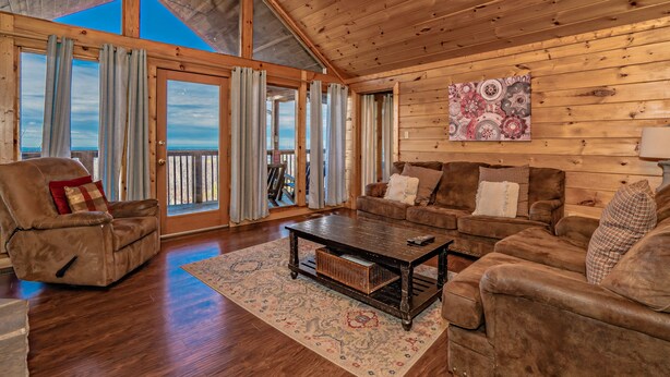 A spacious living room with wooden walls and a large window, furnished with a sofa, armchair, and coffee table. - 🌄 A Great Escape 🍃 Mountain Views, Resort Pool, Hot Tub & Game Room (Sevierville)
