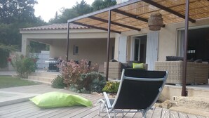 A covered outdoor seating area with a metal chair and a green cushion, a patio with wooden flooring, and a house with a tiled roof and a garden with shrubs and flowers.