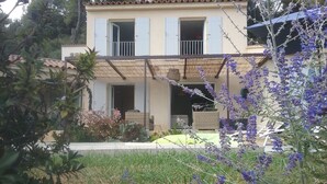 A two-story house with a balcony, a garden with purple flowers, and a small outdoor seating area.