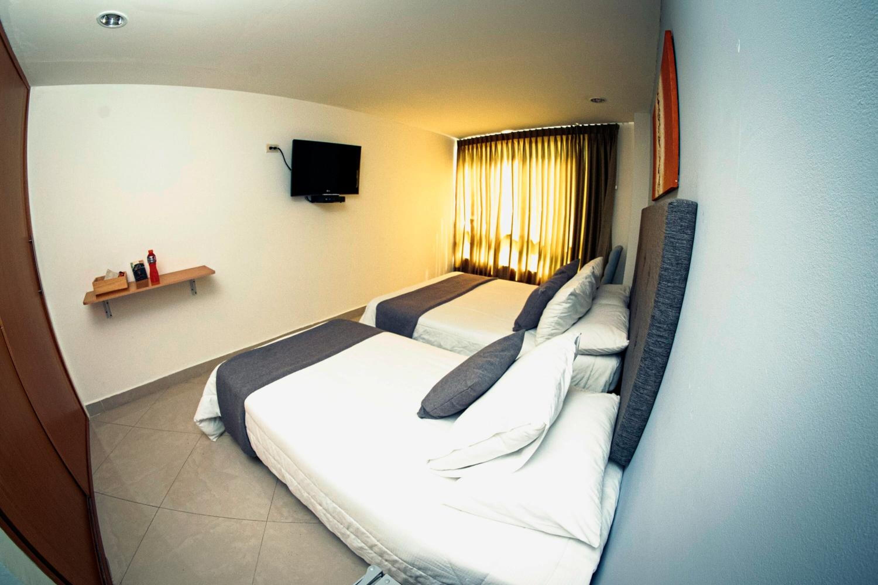 Habitación de hotel con dos camas, un televisor montado en la pared, un estante con artículos de aseo y una ventana con cortinas.