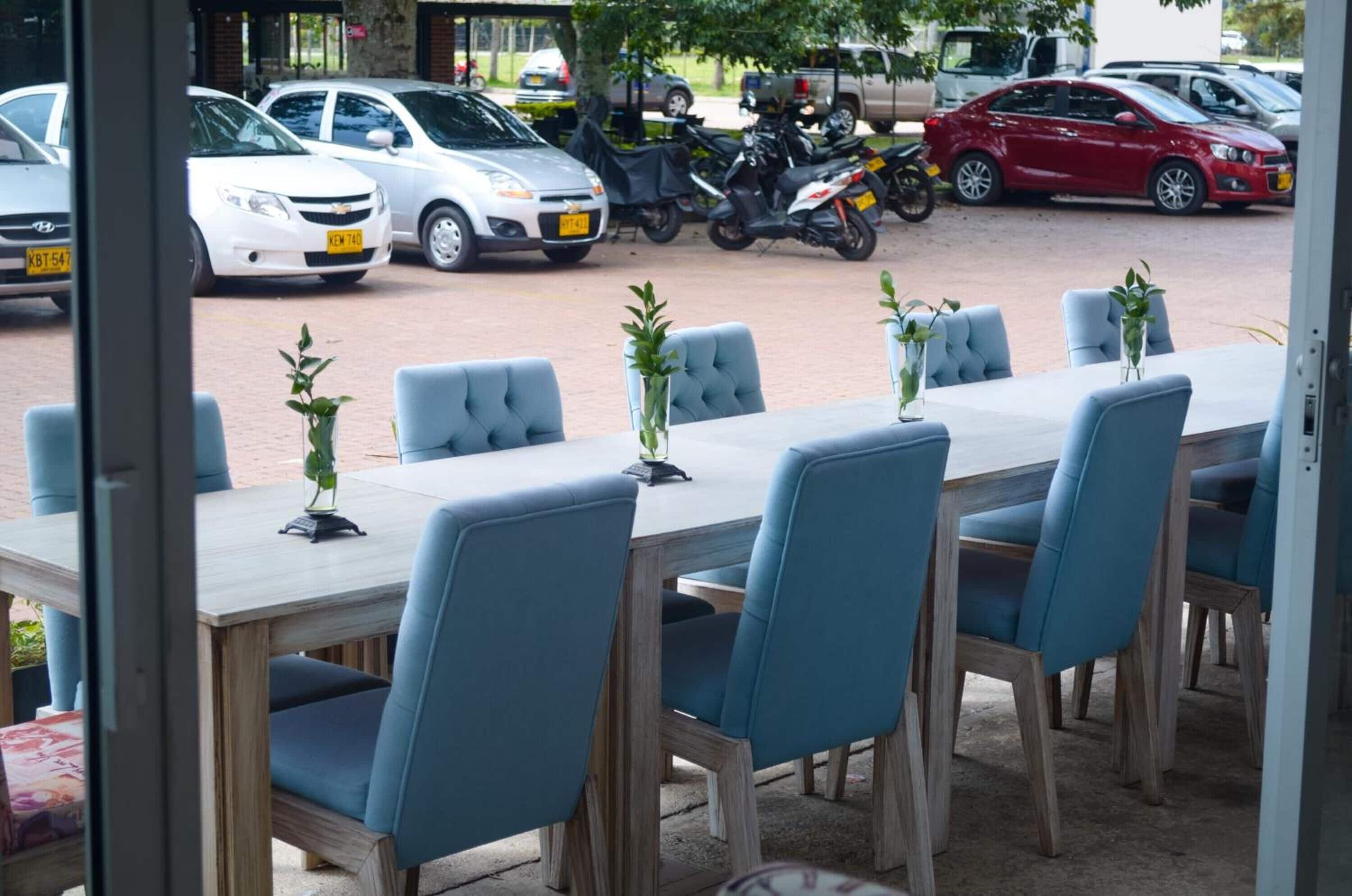 An outdoor dining area with a long table and blue chairs, surrounded by parked cars and motorcycles.