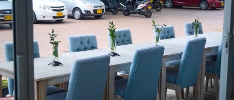 An outdoor dining area with a long table and blue chairs, surrounded by parked cars and motorcycles.
