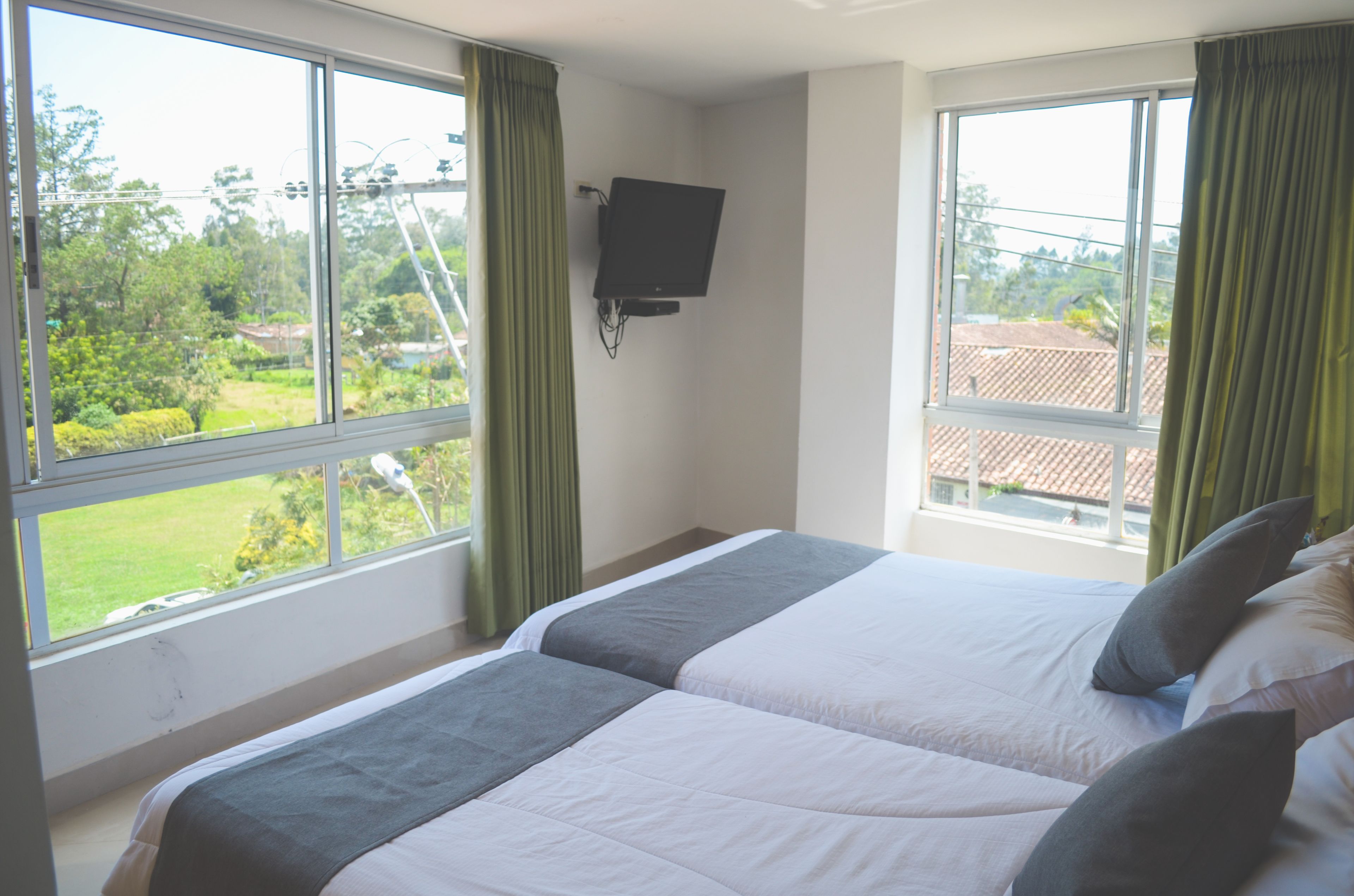 Un dormitorio con dos camas, un televisor montado en la pared y ventanas amplias con cortinas.