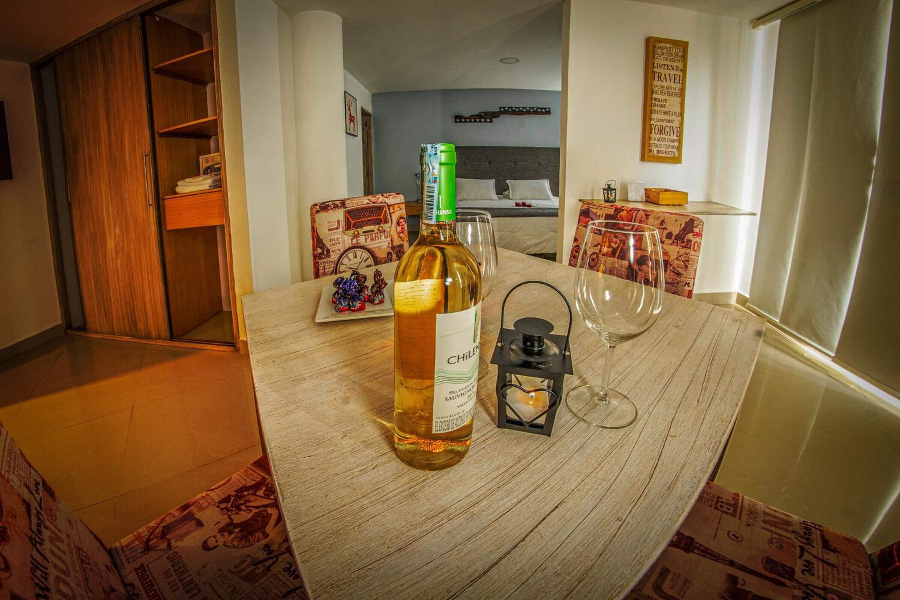 A hotel room with a wooden table, a bottle of wine, a glass, and a lantern. There is a bed with a headboard and a wall with a poster.