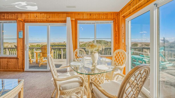 A room with a glass table and chairs, a vase with flowers, and large windows offering a view of the beach and dunes. - Oceanfront Dog Friendly Cottage in Avon with Grill and Screened Porch (Avon)