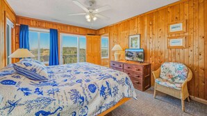 A bedroom with wooden walls, a bed with a blue and white nautical-themed bedspread, a ceiling fan, a dresser, a chair, and a television. - Oceanfront Dog Friendly Cottage in Avon with Grill and Screened Porch (Avon)
