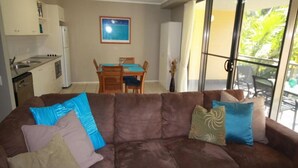 A living room with a brown sofa, a dining table with chairs, a kitchen area, and a large window with a view of greenery. - Baden 90 - Rainbow Shores. Resort Unit - Pools - Tennis Court - Walk To Beach (RAINBOW BEACH)