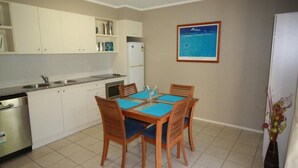 A compact kitchen with white cabinets, a stainless steel refrigerator, and a dining table with blue placemats and wooden chairs. - Baden 90 - Rainbow Shores. Resort Unit - Pools - Tennis Court - Walk To Beach (RAINBOW BEACH)