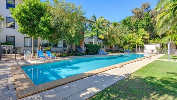 A swimming pool with lounge chairs, surrounded by greenery and buildings. - Baden 90 - Rainbow Shores. Resort Unit - Pools - Tennis Court - Walk To Beach (RAINBOW BEACH)