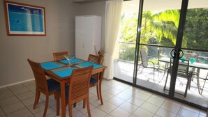 A dining area with a table and chairs, a refrigerator, and a view of a garden through large windows. - Baden 90 - Rainbow Shores. Resort Unit - Pools - Tennis Court - Walk To Beach (RAINBOW BEACH)