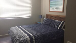 A bedroom with a bed, a nightstand, a lamp, and a window with blinds. - Baden 90 - Rainbow Shores. Resort Unit - Pools - Tennis Court - Walk To Beach (RAINBOW BEACH)