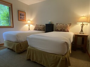 A hotel room with two beds, each with a matching headboard, a nightstand with a lamp, a framed picture on the wall, and a window with a view of greenery.