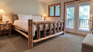 A rustic wooden bed with a simple headboard, a bedside table with a lamp, and a view of the outdoors through a door.