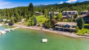 An aerial view of a lakeside property with multiple buildings, a dock with boats, and a well-maintained lawn.