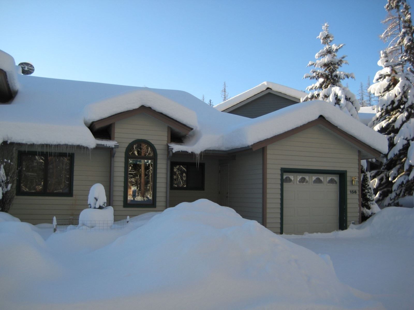 Une maison dont le toit est recouvert de neige, avec un garage et un grand amas de neige devant.
