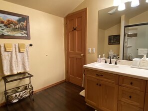 A bathroom with wooden cabinets, a white sink, and a large mirror. There is a painting on the wall and a small table with towels.