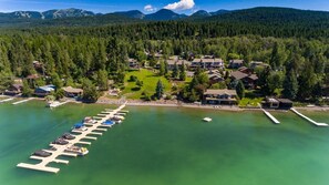 An aerial view of a lakeside resort with a marina, houses, and forested hills in the background. - Mountain Harbor #143 3 Bedroom Condo (Whitefish)