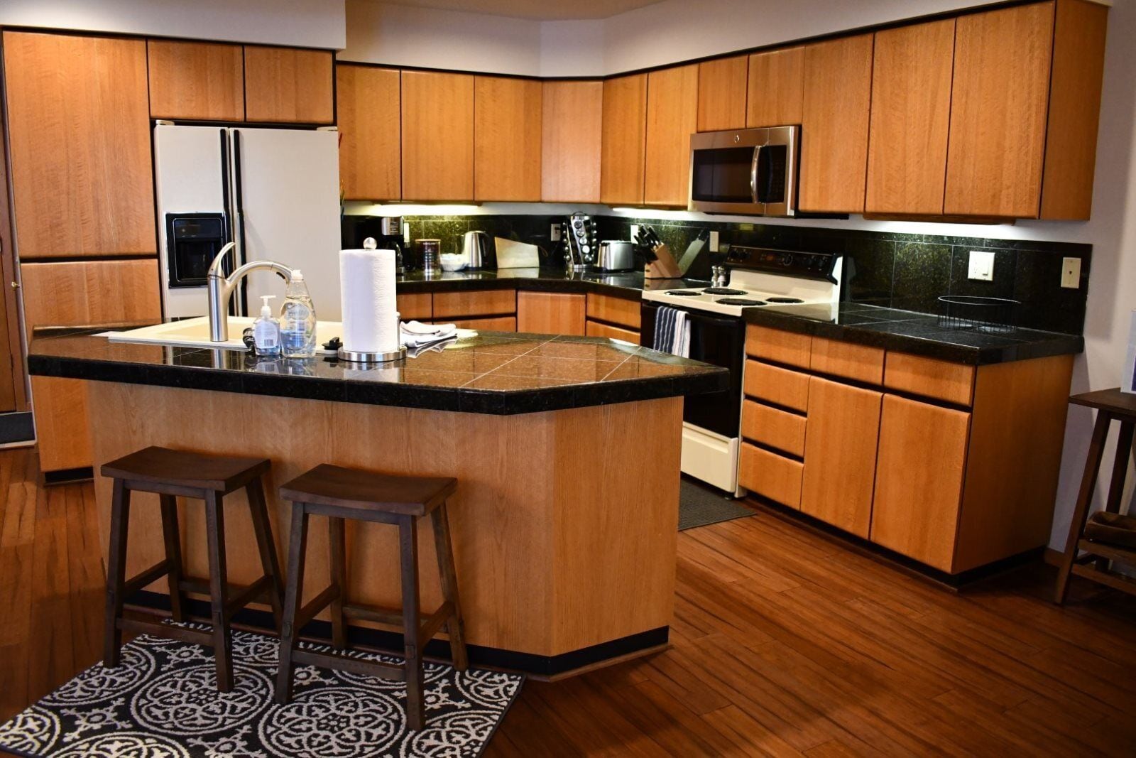 A modern kitchen with wooden cabinets, a black countertop, and a stainless steel microwave. There are two stools at the island.