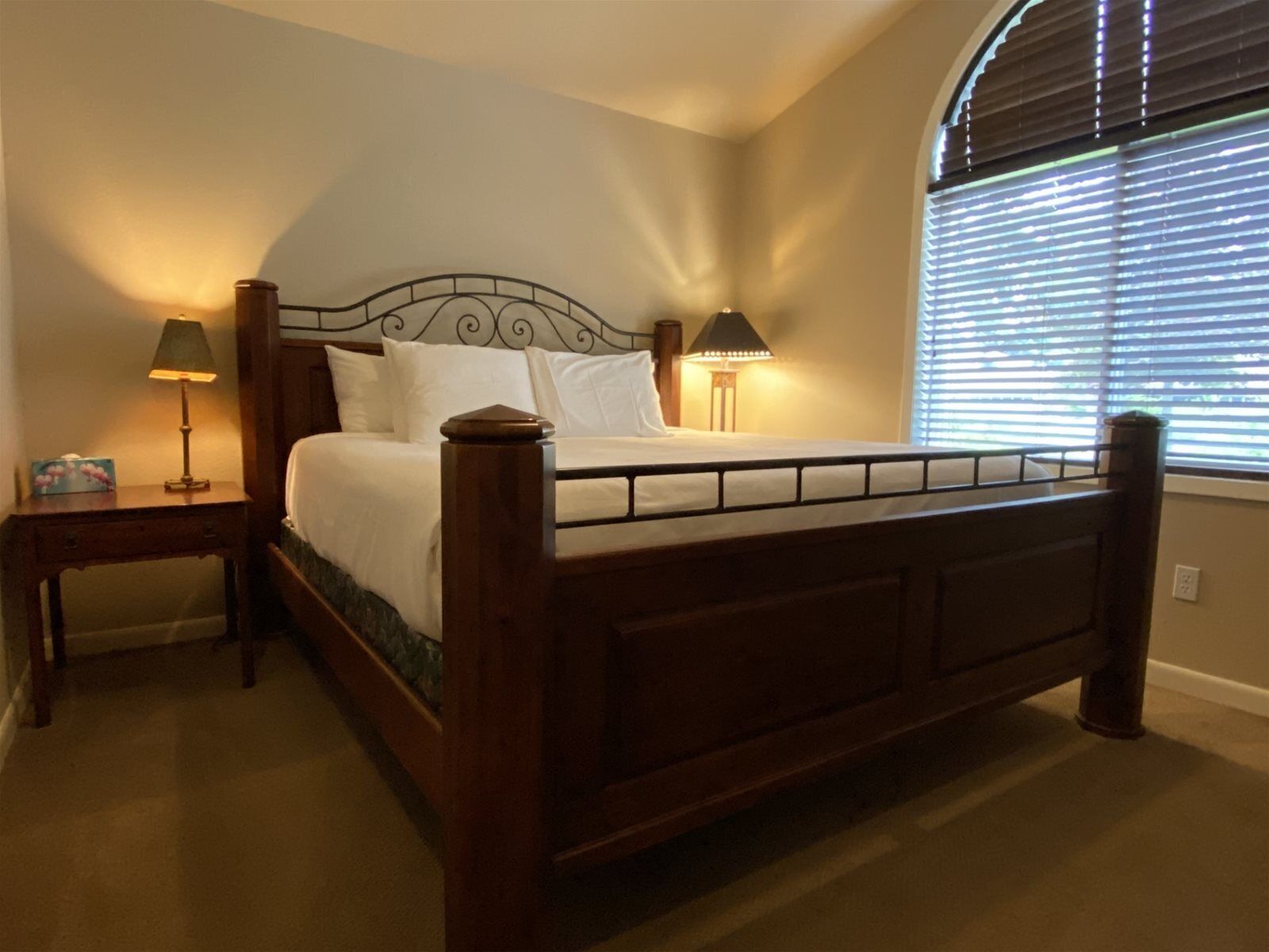 A bedroom with a wooden bed, two bedside tables with lamps, a nightstand, and a window with blinds.