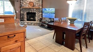 A living room with a stone fireplace, a television, a dining table with chairs, and a kitchen area with wooden cabinets. - Mountain Harbor #127 2 Bedroom Condo (Whitefish)