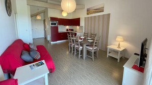 A modern living room with a red sofa, a dining table with chairs, a kitchen area with red cabinets, and a striped curtain. - Joli Appartement avec terrasse face à l'océan (016) (Biscarrosse)