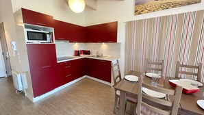 A compact kitchen with red cabinets, a microwave, and a dining area with striped curtains. - Joli Appartement avec terrasse face à l'océan (016) (Biscarrosse)